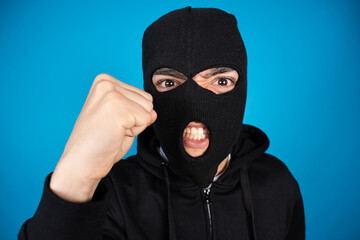 Fototapeta premium Angry criminal menacing fist up to the camera isolated on blue background. Delinquent wearing face mask.