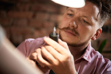 Concentrated male tattooist with beard and moustache looking at tattoo gun attentively under lamp light