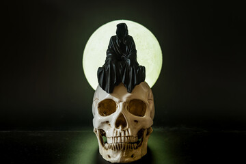 A statue of a grim reaper sitting on a skull on a full moon background.