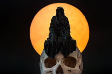 A statue of a grim reaper sitting on a skull on a full moon background.