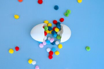 Multi-colored pom-poms falling out of an egg broken into two parts on a blue background, a funny egg