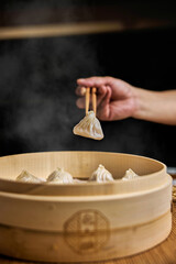 Close up of Chinese steamed Xiaolongbao held in chopsticks, being taken out of bamboo steamer with rustic wood.