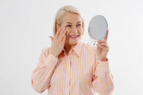 Happy Middle aged Woman looking in mirror on her face with wrinkles on the forehead and wrinkle skin. Collagen and face injections concept. Menopause. Copy space and mock up.