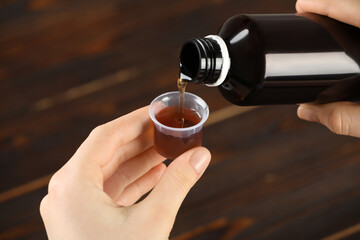 Woman pouring cough syrup into measuring cup at wooden table, closeup