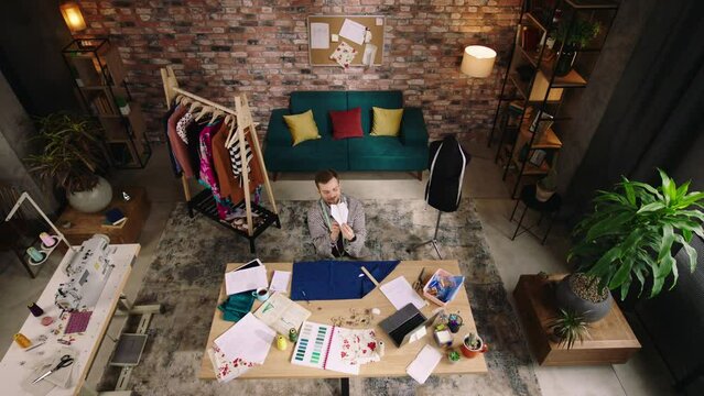 Top view. A man is in a very comfy and well decorated home office, he is playing around with an airplane while sitting at his desk and continuing on with his designer work on a piece of blue fabric