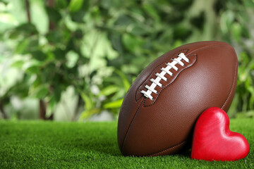American football ball and red heart on green grass against blurred background. Space for text