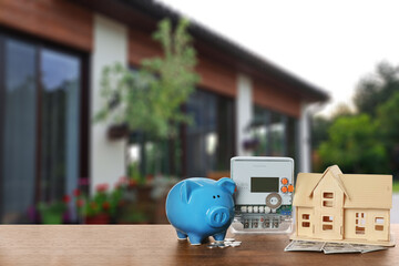Electricity meter, house model and piggy bank on wooden table against against blurred view of...