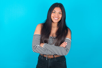 young latin woman wearing striped T-shirt over blue background being happy smiling and crossed arms looking confident at the camera. Positive and confident person.