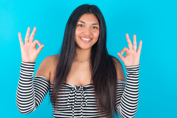 Glad young latin woman wearing striped T-shirt over blue background shows ok sign with both hands as expresses approval, has cheerful expression, being optimistic.