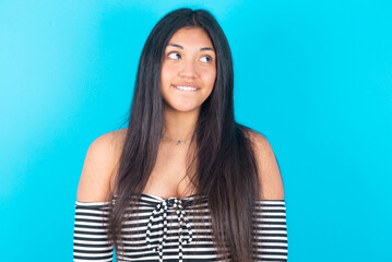 Fototapeta premium young latin woman wearing striped T-shirt over blue background with thoughtful expression, looks away keeps hands down bitting his lip thinks about something pleasant.