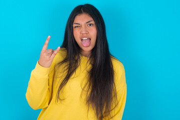 Portrait of a crazy young latin woman wearing yellow sweater over blue background showing tongue horns up gesture, expressing excitement of being on concert of band.
