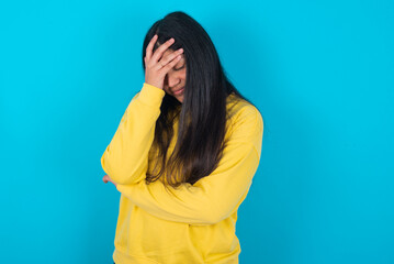 young latin woman wearing yellow sweater over blue background making facepalm gesture while smiling...