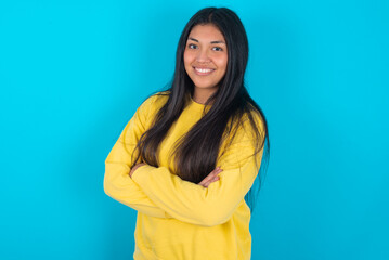 Fototapeta premium Image of cheerful young latin woman wearing yellow sweater over blue background with arms crossed. Looking and smiling at the camera. Confidence concept.