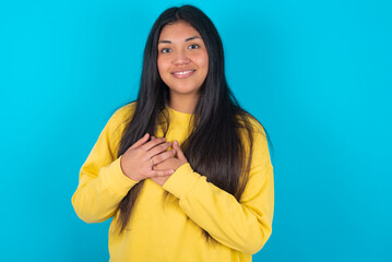 Honest young latin woman wearing yellow sweater over blue background keeps hands on chest, touched...