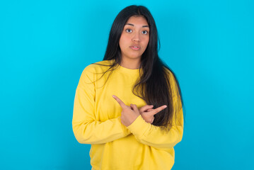 Confused young latin woman wearing yellow sweater over blue background chooses between two ways, points at both sides with crossed hands, feels doubt. Need your advice.
