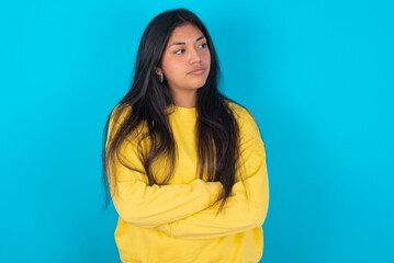 Image of upset young latin woman wearing yellow sweater over blue background with arms crossed. Looking with disappointed expression aside after listening to bad news.