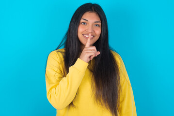 Smiling young latin woman wearing yellow sweater over blue background makes shush gesture, holds...