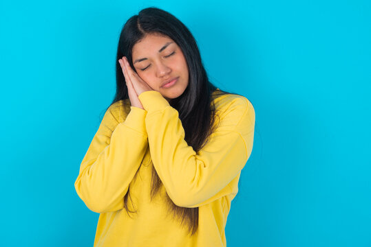 Relax and sleep time. Tired young latin woman wearing yellow sweater over blue background with closed eyes leaning on palms making sleeping gesture.