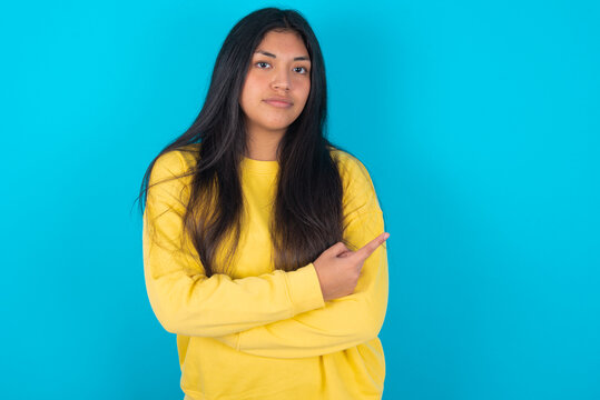 young latin woman wearing yellow sweater over blue background smiling broadly at camera, pointing fingers away, showing something interesting and exciting.