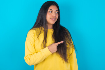 Fototapeta premium Happy cheerful smiling young latin woman wearing yellow sweater over blue background looking and pointing aside with hand. Copy space and advertisement concept.
