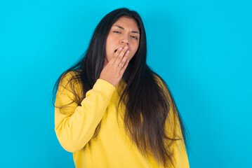 Sleepy young latin woman wearing yellow sweater over blue background yawning with messy hair, feeling tired after sleepless night, yawning, covering mouth with palm.