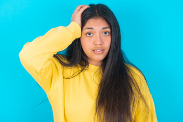 Portrait of confused young latin woman wearing yellow sweater over blue background holding hand on hair and frowning, panicking, losing memory. Worried and anxious can not remember anything.