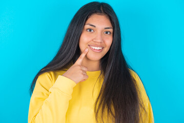 Obraz premium Carefree successful young latin woman wearing yellow sweater over blue background touching jawline gazing camera tilting head grinning white teeth delighted. Dental care concept.