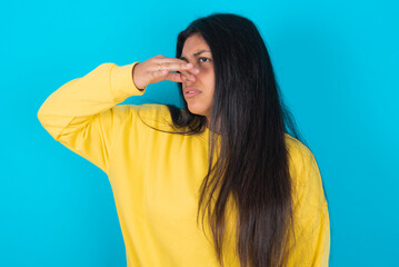 Displeased young latin woman wearing yellow sweater over blue background plugs nose as smells something stink and unpleasant, feels aversion, hates disgusting scent. - obrazy, fototapety, plakaty