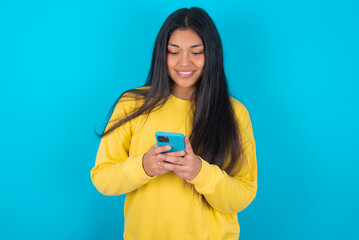 Smiling young latin woman wearing yellow sweater over blue background using cell phone, messaging,...
