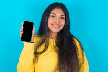 Smiling young latin woman wearing yellow sweater over blue background showing  empty phone screen. Advertisement and communication concept.