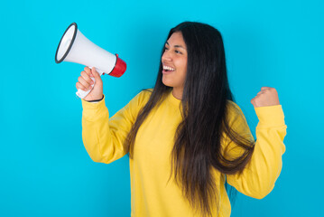 young latin woman wearing yellow sweater over blue background communicates shouting loud holding a megaphone, expressing success and positive concept, idea for marketing or sales.