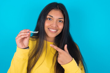 young latin woman wearing yellow sweater over blue background holding an invisible aligner and...