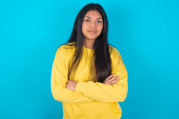 Obraz premium Self confident serious calm young latin woman wearing yellow sweater over blue background stands with arms folded. Shows professional vibe stands in assertive pose.