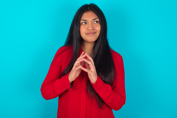 young latin woman wearing red shirt over blue background steepled fingers and looks mysterious aside has great evil plan in mind