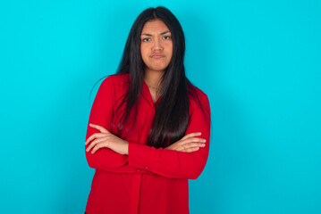 Gloomy dissatisfied young latin woman wearing red shirt over blue background looks with miserable expression at camera from under forehead, makes unhappy grimace