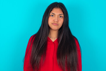 Offended dissatisfied young latin woman wearing red shirt over blue background with moody displeased expression at camera being disappointed by something