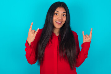 young latin woman wearing red shirt over blue background makes rock n roll sign looks self...