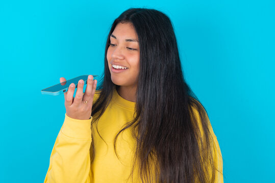 Smiling young latin woman wearing yellow sweater over blue background sending voice message on her smart phone. Communication and new technologies concept.