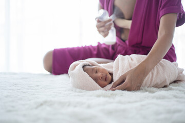Newborn baby 27 days old wrap in blanket on a bed while mother pumping milk to bottles in blurred background, selective focus