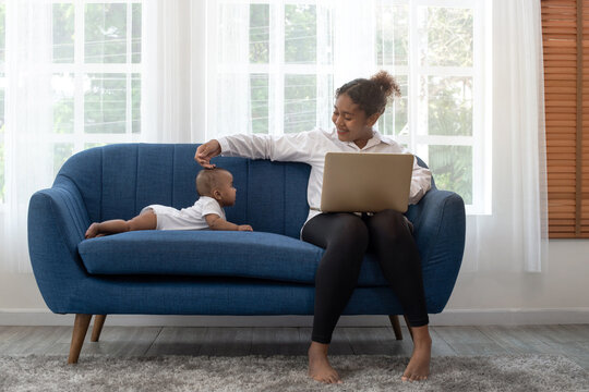 African mother working remotely on laptop while taking care of her baby on couch, freelance single mom working at home, 5 months baby old lying on couch near her