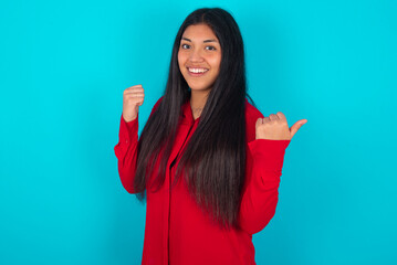 Hooray cool young latin woman wearing red shirt over blue background point back empty space hand fist