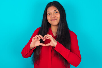 Serious young latin woman wearing red shirt over blue background keeps hands crossed stands in thoughtful pose concentrated somewhere