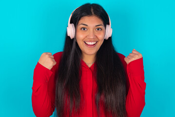 Emotional young latin woman wearing red shirt over exclaims loudly feels like winner raises...