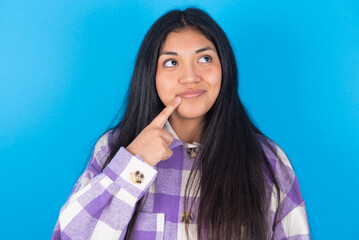 Fototapeta premium Lovely dreamy young latin woman wearing plaid shirt over blue background keeps finger near lips looks aside copy space.