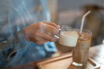 A hand pouring a glass of milk cream into iced latte coffee on a wooden bar over a cafe glass window reflex at a Cafe coffee shop. Cold brew refreshment summer drink with copy space. Selective focus
