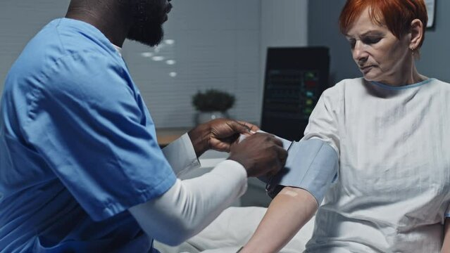 Tilting up and down of young African American male nurse wearing blue scrub suit sitting on bed in hospital ward, measuring blood pressure of mature red-haired Caucasian woman