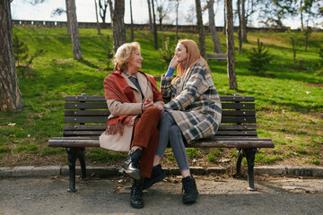 A young ginger woman is sitting with her grandmother and talking on the park bench in the cold weather.