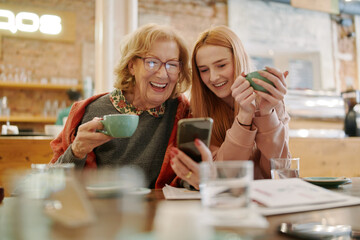 Happy grandmother and her adolescent granddaughter sitting in a cafe, enjoying coffee and using smart phone for funny videos. - 501709662