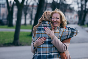 A grandmother is hugging her adult granddaughter in the park. They are greeting, and happy to see each other.