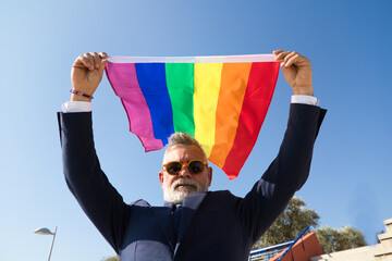 Mature gay man, executive, grey-haired, bearded, sunglasses, jacket and tie, waving the new lgbtiq+...